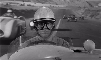 Movie still from “On the Beach” (1959), directed by Stanley Kramer – A man wearing a helmet and glasses driving a car; Close Up shot, Over the shoulder angle