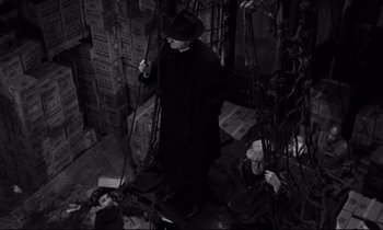 Movie still from “On the Waterfront” (1954), directed by Elia Kazan – A man in a black coat and a black hat standing in a room; Wide shot, Overhead angle