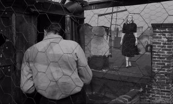 Movie still from “On the Waterfront” (1954), directed by Elia Kazan – A man and a woman are standing behind a chain link fence; Wide shot, Low angle
