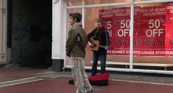 Movie still from “Once” (2007), directed by John Carney – A man playing a guitar while another man smokes a cigarette on the sidewalk; Wide shot, Over the shoulder angle