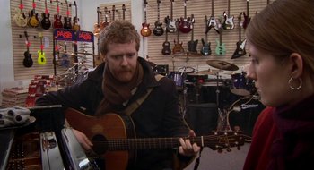 Movie still from “Once” (2007), directed by John Carney – A man playing an acoustic guitar in front of a wall of guitars; Medium shot, Over the shoulder angle