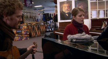 Movie still from “Once” (2007), directed by John Carney – A woman is playing a guitar in a music store; Medium shot, Over the shoulder angle