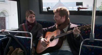 Movie still from “Once” (2007), directed by John Carney – A man sitting on top of a couch playing a guitar; Medium shot, High angle