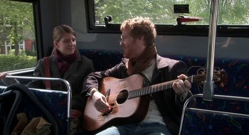 Movie still from “Once” (2007), directed by John Carney – A man sitting on top of a couch playing a guitar; Medium shot, Over the shoulder angle