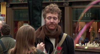 Movie still from “Once” (2007), directed by John Carney – A man wearing a brown scarf and holding a red rose; Medium shot, Over the shoulder angle