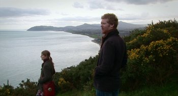 Movie still from “Once” (2007), directed by John Carney – A man and a woman standing on top of a hill near a body of water; Medium shot, Over the shoulder angle