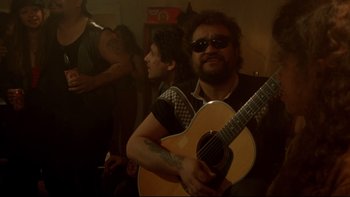 Movie still from “Once Were Warriors” (1994), directed by Lee Tamahori – A man holding a guitar in front of other people; Medium shot, Over the shoulder angle
