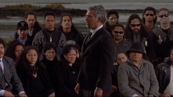 Movie still from “Once Were Warriors” (1994), directed by Lee Tamahori – A man in a suit and tie standing in front of a group of people; Medium shot, Over the shoulder angle