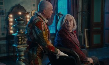 Movie still from “One Hundred and One Nights” (1995), directed by Agnès Varda – A man in a clown suit and an older woman in a chair; Medium shot, Over the shoulder angle
