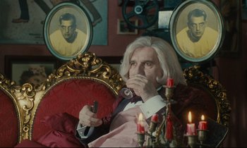 Movie still from “One Hundred and One Nights” (1995), directed by Agnès Varda – An older man sitting in a chair with a remote control in his hand; Medium shot, Over the shoulder angle