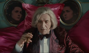 Movie still from “One Hundred and One Nights” (1995), directed by Agnès Varda – An older man sitting in front of a painting; Medium shot, Over the shoulder angle