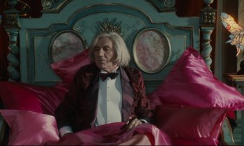 Movie still from “One Hundred and One Nights” (1995), directed by Agnès Varda – An old man is sitting on a bed wearing a crown; Medium shot, Over the shoulder angle