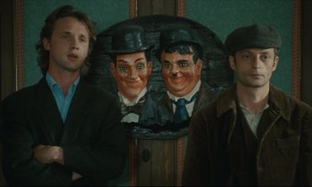 Movie still from “One Hundred and One Nights” (1995), directed by Agnès Varda – Three men standing next to a painting of laurel and hardy; Medium shot, Over the shoulder angle