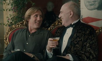 Movie still from “One Hundred and One Nights” (1995), directed by Agnès Varda – Two men are sitting and laughing while holding a drink; Medium shot, Over the shoulder angle