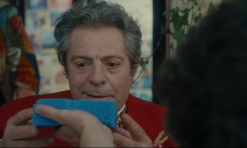 Movie still from “One Hundred and One Nights” (1995), directed by Agnès Varda – An older man in a red jacket holding a blue sponge; Close Up shot, Over the shoulder angle