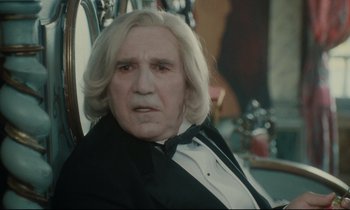 Movie still from “One Hundred and One Nights” (1995), directed by Agnès Varda – A person sitting in a chair wearing a suit; Close Up shot, Over the shoulder angle