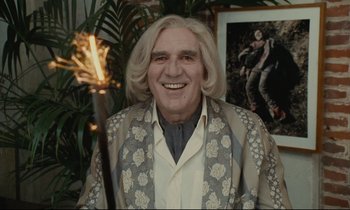 Movie still from “One Hundred and One Nights” (1995), directed by Agnès Varda – An older man holding a sparkler and smiling for the camera; Medium shot, Low angle