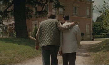 Movie still from “One Hundred and One Nights” (1995), directed by Agnès Varda – Two men walking down a dirt road near a building; Wide shot, Over the shoulder angle