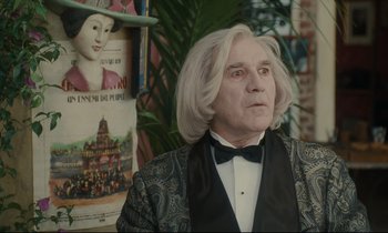 Movie still from “One Hundred and One Nights” (1995), directed by Agnès Varda – A man with white hair wearing a suit and bow tie in front of a poster; Medium shot, Over the shoulder angle