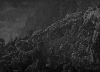 Movie still from “One Million B.C.” (1940), directed by Hal Roach Jr. – A black and white photo of a group of people sitting on rocks; Extreme Wide shot, High angle
