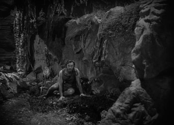 Movie still from “One Million B.C.” (1940), directed by Hal Roach Jr. – A man crouching on the ground in front of a rock wall; Wide shot, High angle