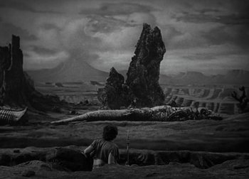 Movie still from “One Million B.C.” (1940), directed by Hal Roach Jr. – A black and white photo of a man sitting in front of a mountain; Extreme Wide shot, High angle