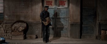 Movie still from “One Second” (2020), directed by Yimou Zhang – A man standing in front of a building holding a bag; Wide shot, High angle