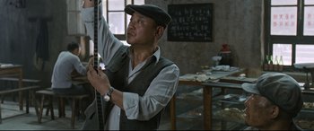Movie still from “One Second” (2020), directed by Yimou Zhang – A man in a hat and vest holding a ruler; Medium shot, Low angle