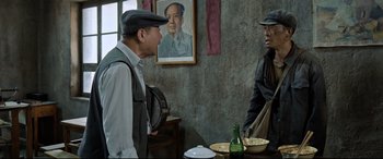 Movie still from “One Second” (2020), directed by Yimou Zhang – Two men are standing at a table in front of a portrait of mao zedong on the wall; Medium shot, Over the shoulder angle