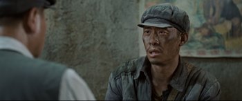 Movie still from “One Second” (2020), directed by Yimou Zhang – A man wearing a hat sitting in front of a stone wall; Close Up shot, Over the shoulder angle