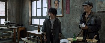 Movie still from “One Second” (2020), directed by Yimou Zhang – A man standing at a table in front of a portrait of mao zedong on the wall; Medium shot, Over the shoulder angle