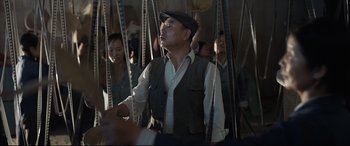 Movie still from “One Second” (2020), directed by Yimou Zhang – A man in a hat and vest standing in front of a group of people; Medium shot, Low angle