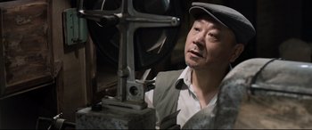 Movie still from “One Second” (2020), directed by Yimou Zhang – A man in a hat is working on a machine; Close Up shot, Low angle