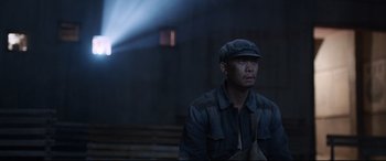 Movie still from “One Second” (2020), directed by Yimou Zhang – A man wearing a hat and jacket standing in front of a building; Medium shot, Over the shoulder angle