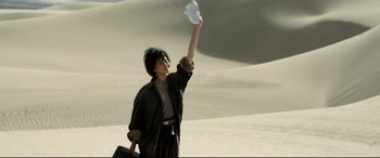 Movie still from “One Second” (2020), directed by Yimou Zhang – A person standing in the sand holding a paper in the air; Medium shot, Low angle