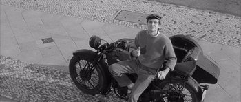 Movie still from “One, Two, Three” (1961), directed by Billy Wilder – A man sitting on the back of a parked motorcycle; Medium shot, High angle