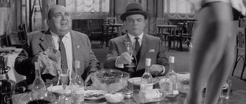 Movie still from “One, Two, Three” (1961), directed by Billy Wilder – A man in a suit and hat sitting at a table with a glass of wine; Medium shot, High angle