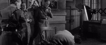 Movie still from “One, Two, Three” (1961), directed by Billy Wilder – An old photo of a man in a military uniform smoking a cigarette in front of a chair; Medium shot, Over the shoulder angle