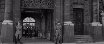 Movie still from “One, Two, Three” (1961), directed by Billy Wilder – An old photo of a man in a uniform standing in front of a building; Wide shot, Low angle
