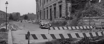 Movie still from “One, Two, Three” (1961), directed by Billy Wilder – An old car driving down the street near a building; Wide shot, Low angle