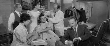 Movie still from “One, Two, Three” (1961), directed by Billy Wilder – A group of people in a room with a man cutting another man's hair; Medium shot, Over the shoulder angle
