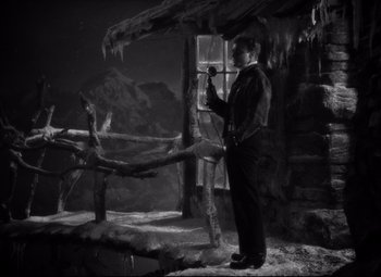 Movie still from “Only Angels Have Wings” (1939), directed by Howard Hawks – A black and white photo of a man standing in front of a log cabin; Wide shot, High angle