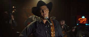 Movie still from “Only the Brave” (2017), directed by Joseph Kosinski – An older man wearing a cowboy hat while holding a microphone; Close Up shot, Over the shoulder angle