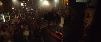 Movie still from “Only the Brave” (2017), directed by Joseph Kosinski – A man in a cowboy hat is holding a guitar in front of a crowd of onlookers; Wide shot, Over the shoulder angle