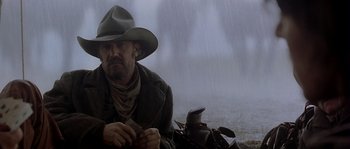 Movie still from “Open Range” (2003), directed by Kevin Costner – A man in a cowboy hat sitting on the ground in the rain; Medium shot, Low angle