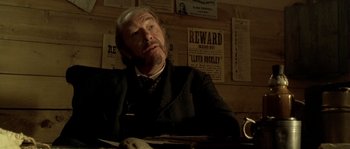 Movie still from “Open Range” (2003), directed by Kevin Costner – An older man sitting at a table in front of a wall of wanted posters; Close Up shot, Over the shoulder angle