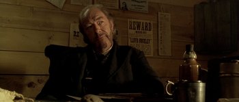 Movie still from “Open Range” (2003), directed by Kevin Costner – An older man sitting at a table in front of a wall of wanted posters; Close Up shot, Over the shoulder angle