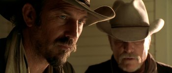 Movie still from “Open Range” (2003), directed by Kevin Costner – A man in a cowboy hat looking at the camera with another man; Close Up shot, Low angle