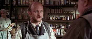 Movie still from “Open Range” (2003), directed by Kevin Costner – A man with a beard wearing an apron in front of shelves of medicine; Close Up shot, Over the shoulder angle
