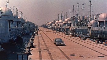 Movie still from “Operation Petticoat” (1959), directed by Blake Edwards – A car driving down a street next to a harbor; Extreme Wide shot, High angle